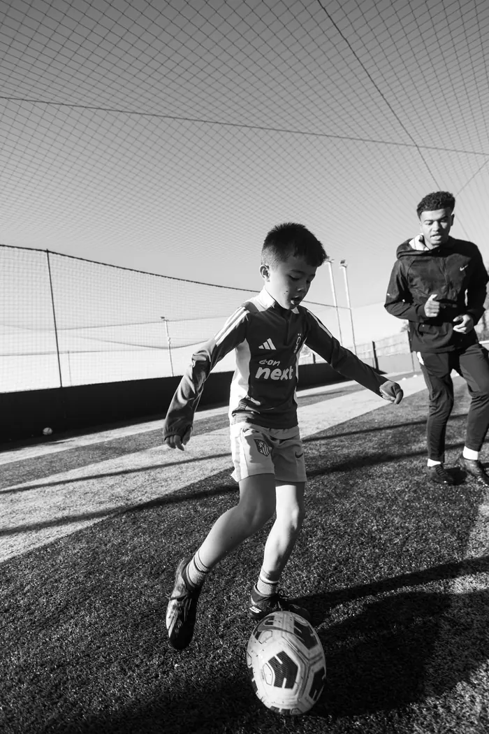 A young player with the ball and the coach close behind, in black and white